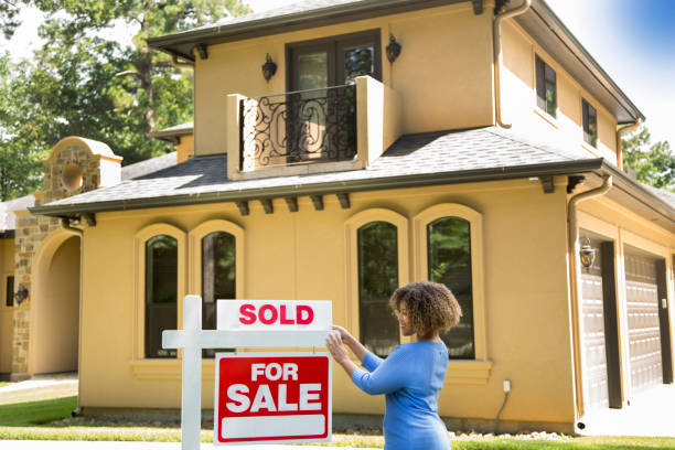 Lovely young adult Real Estate Agent standing beside her for sale sign in front yard of home.  She adds the SOLD sign and wears a blue top and jeans.